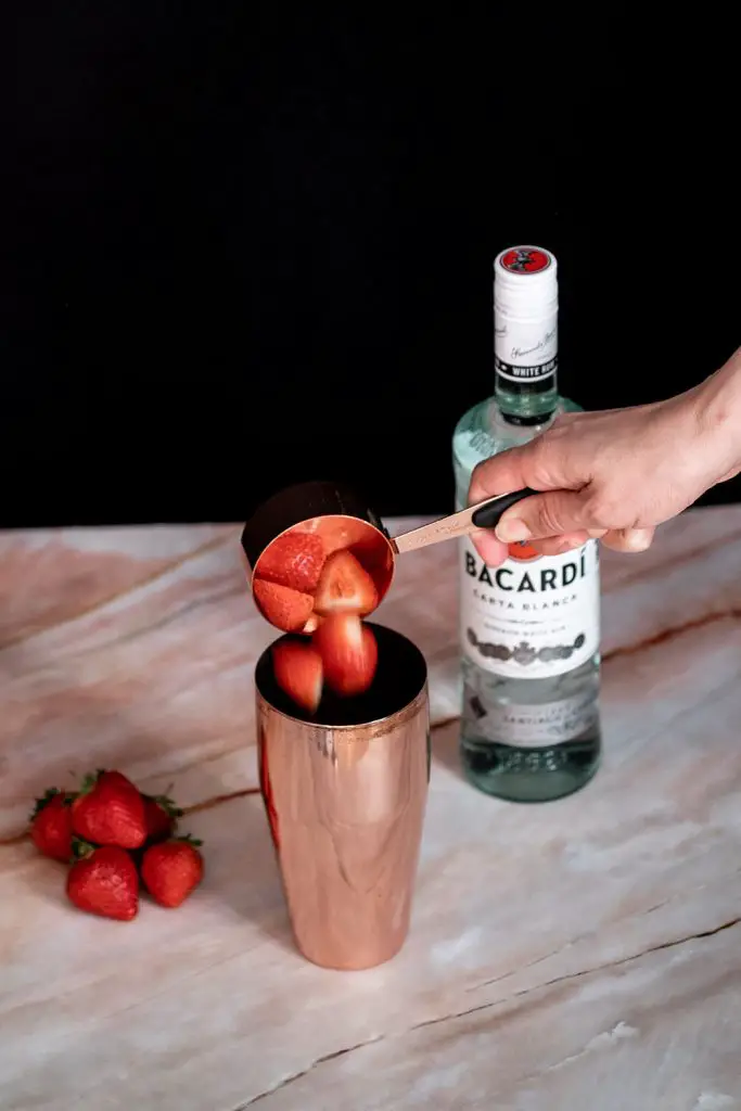 A hand putting halved strawberries in a cocktail shaker.