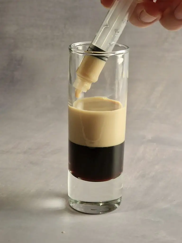 layering a cocktail shot glass by using an injector, pouring baileys on top of coffee liqueur while making b52 cocktail shot. 