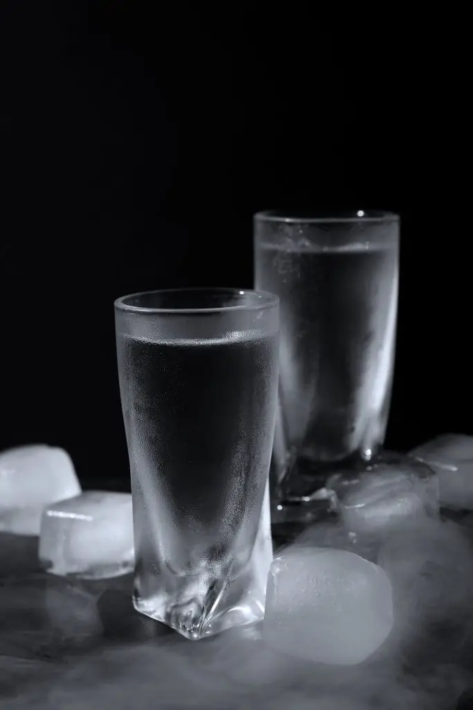 2 chilled, ice cold shot glasses with some ice parts next to them, on a dark background.