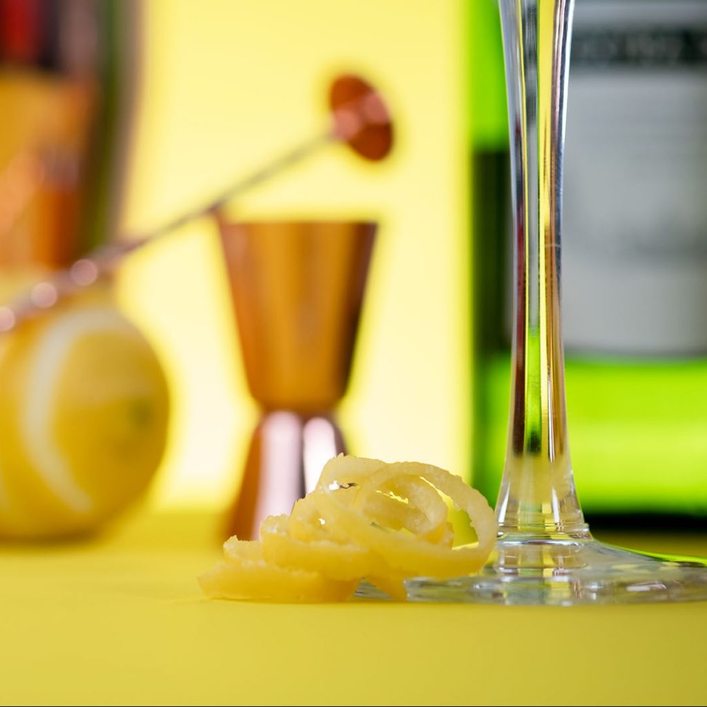 Lemon peek at the bottom of a cocktail glass with a cocktail jigger, lemon and dry vermouth bottle blurry on the background