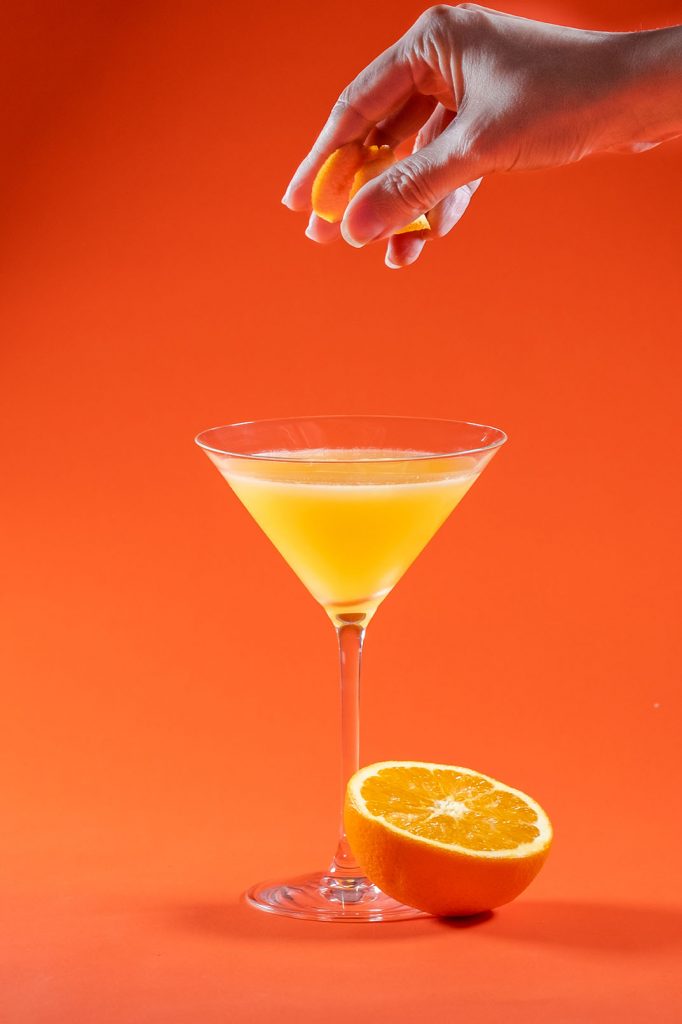 woman hand squeezing orange peel twist to an orange martini with a half sliced orange