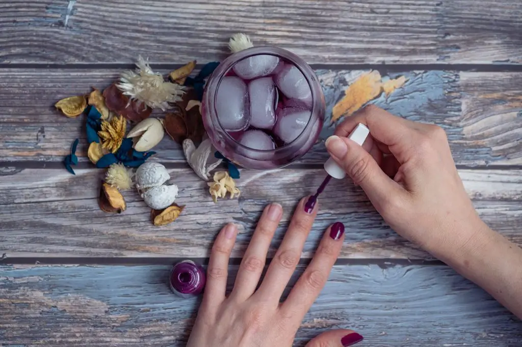 hand painting nails with purple nail polish and a purple cocktail on a wooden table with some dry flowers