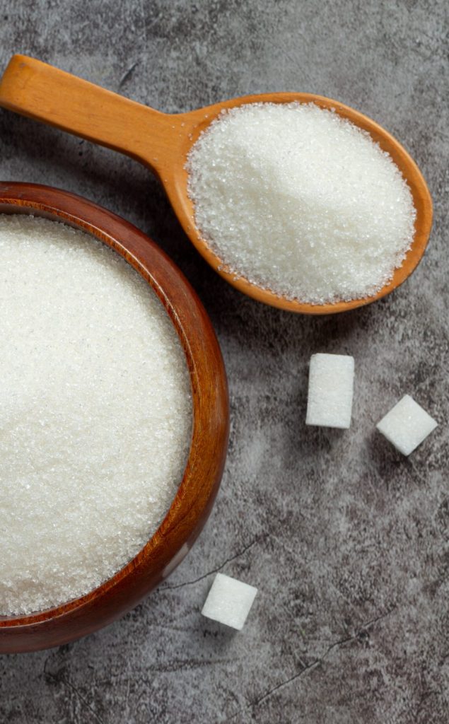 a bowl of caster sugar and a spoon full with sugar with 3 sugar cubes on a concrete looking ground for homemade cocktail ingredients