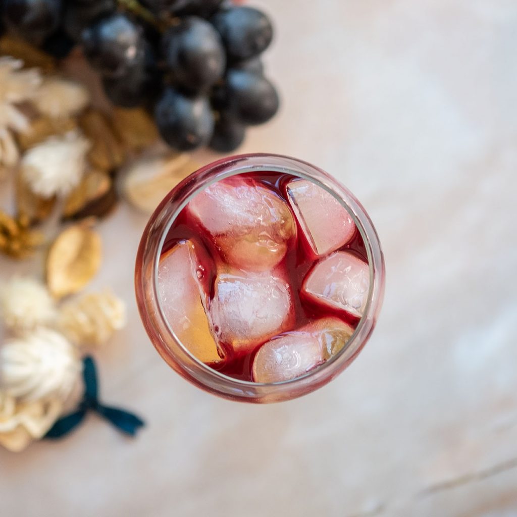 Top view of a cocktail glass full of ice and with red liquid, on a marble background, grapes and dry flowers blurry on the background