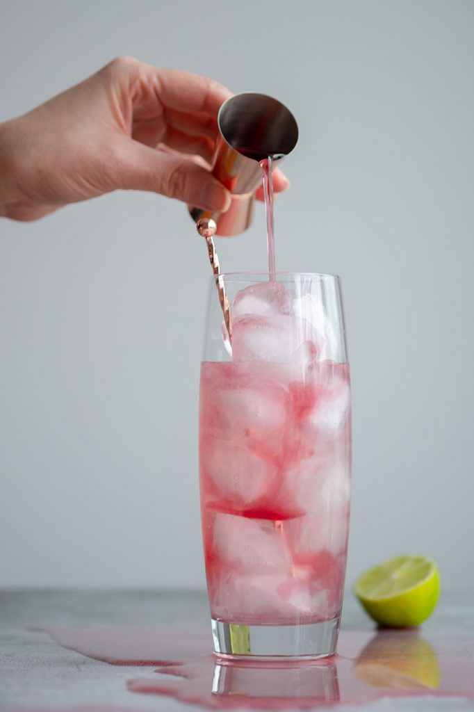 A hand pouring cranberry juice in a tall glass, making a City Rickey Cocktail, filled with ice.