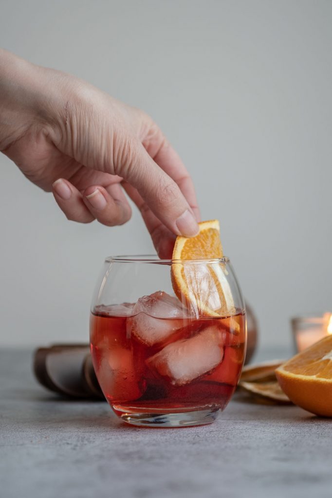 A hand garnishing Negroni with a slice of orange in an old fashioned glass filled with ice 