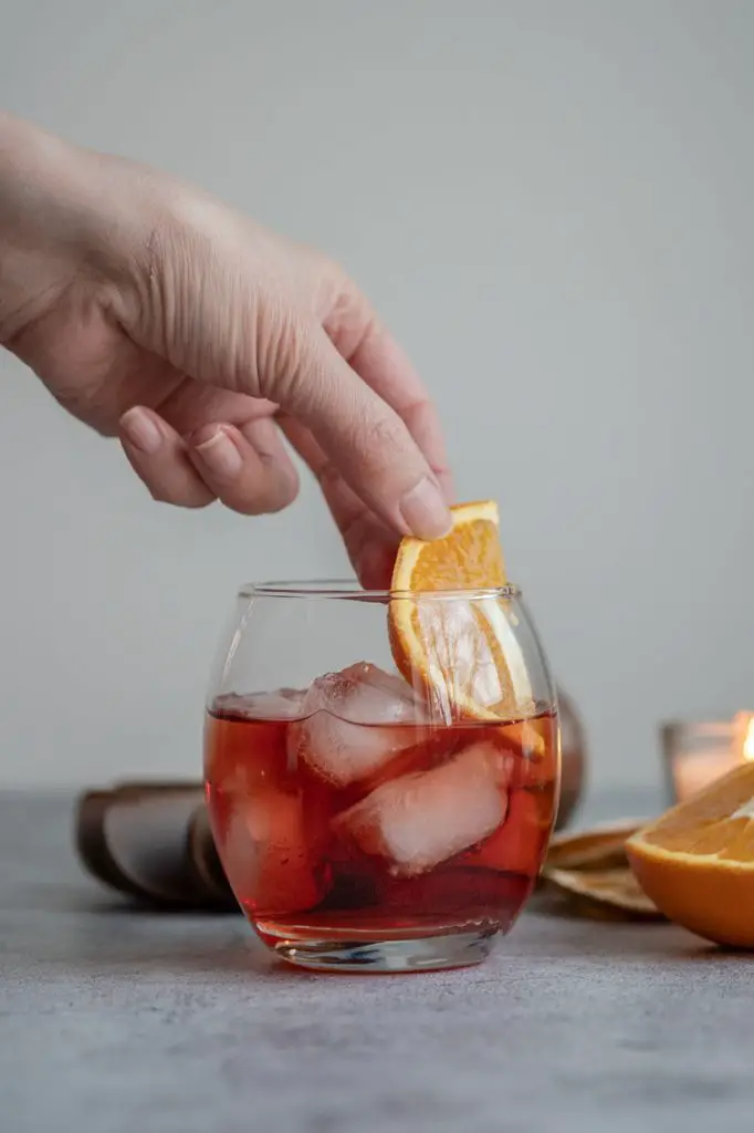 A hand garnishing Negroni with a slice of orange in an old fashioned glass filled with ice