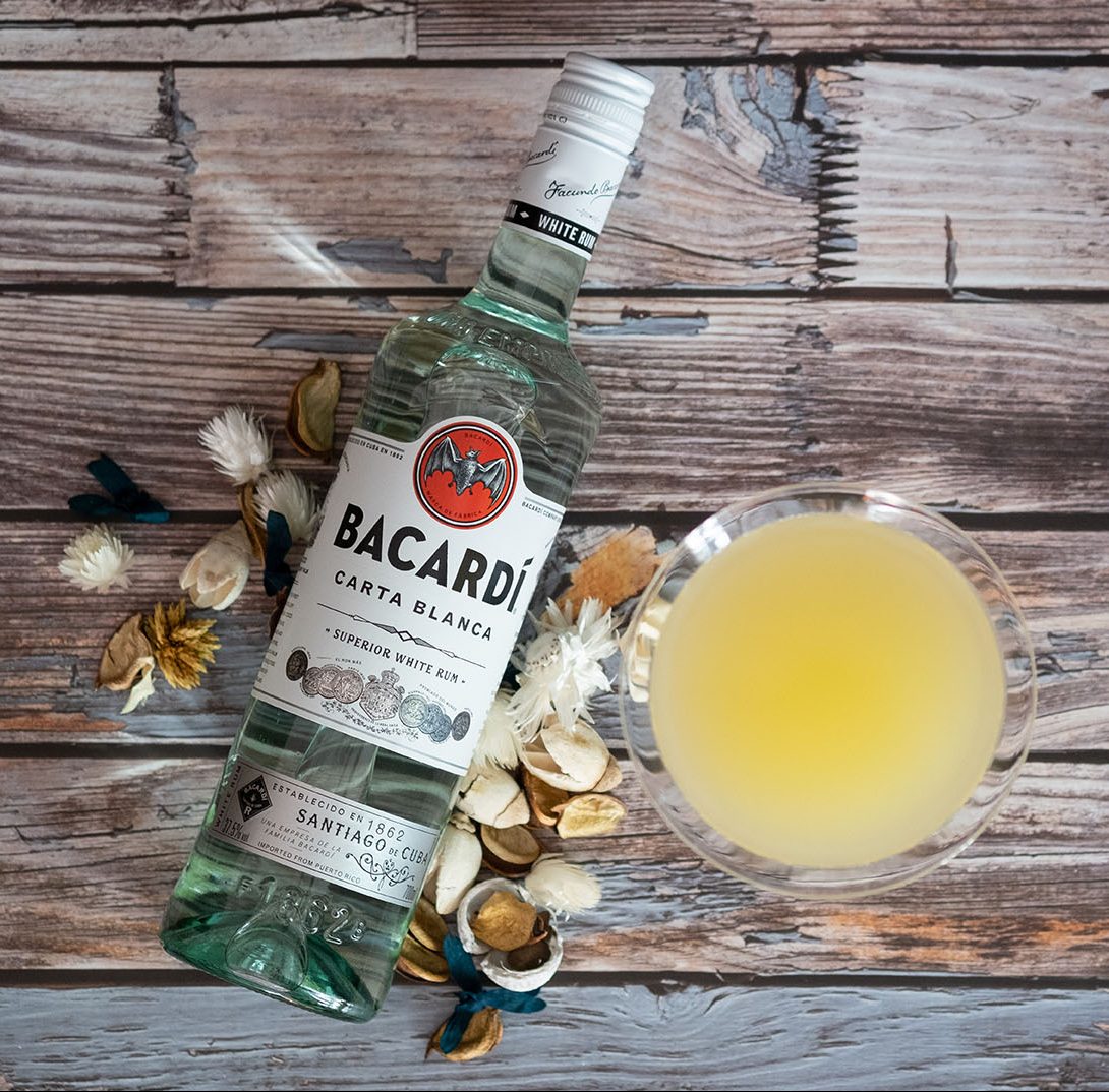 A bottle of bacardi lying on a wooden table with a banana daiquiri cocktail and some dried flowers