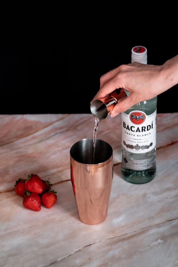 A hand pouring simple syrup in a cocktail shaker to make strawberry daiquiri