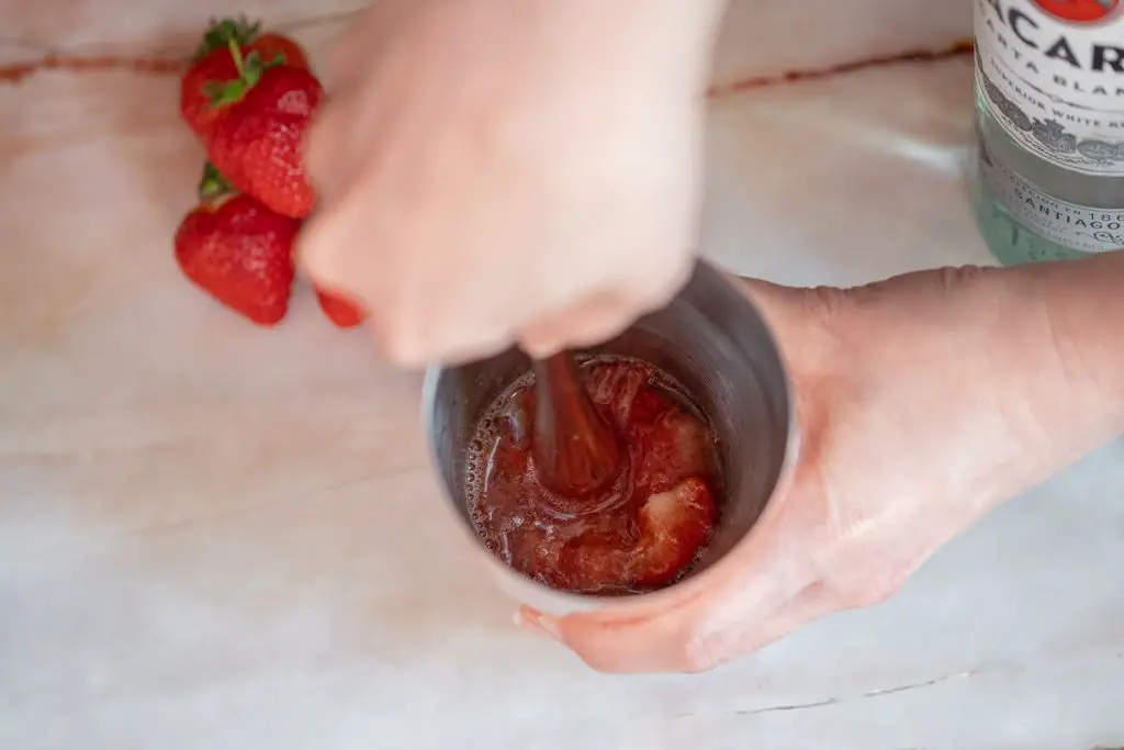 A hand muddling strawberries in a cocktail shaker, some strawberries on the background on a marble table.