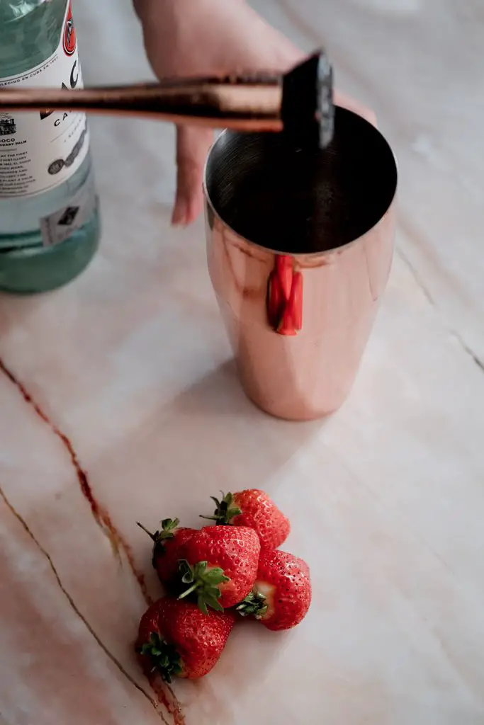 A muddler used for muddling strawberries in a shaker, on a marble looking table with some fresh strawberries.