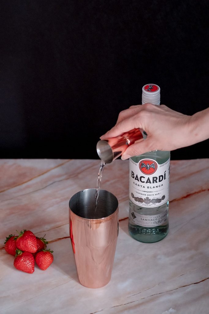 A hand pouring Bacardi branded rum into a cocktail shaker on a marble looking table, with some strawberries and a bottle of Bacardi on it.
