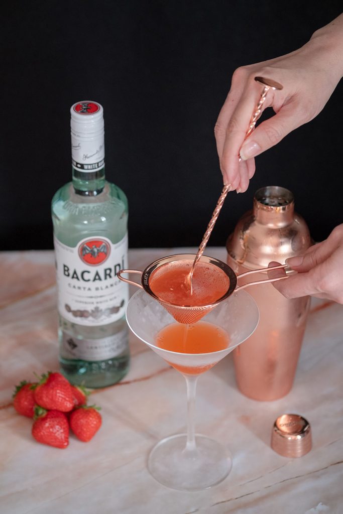 A hand fine straining strawberry daiquiri into a chilled martini glass.
