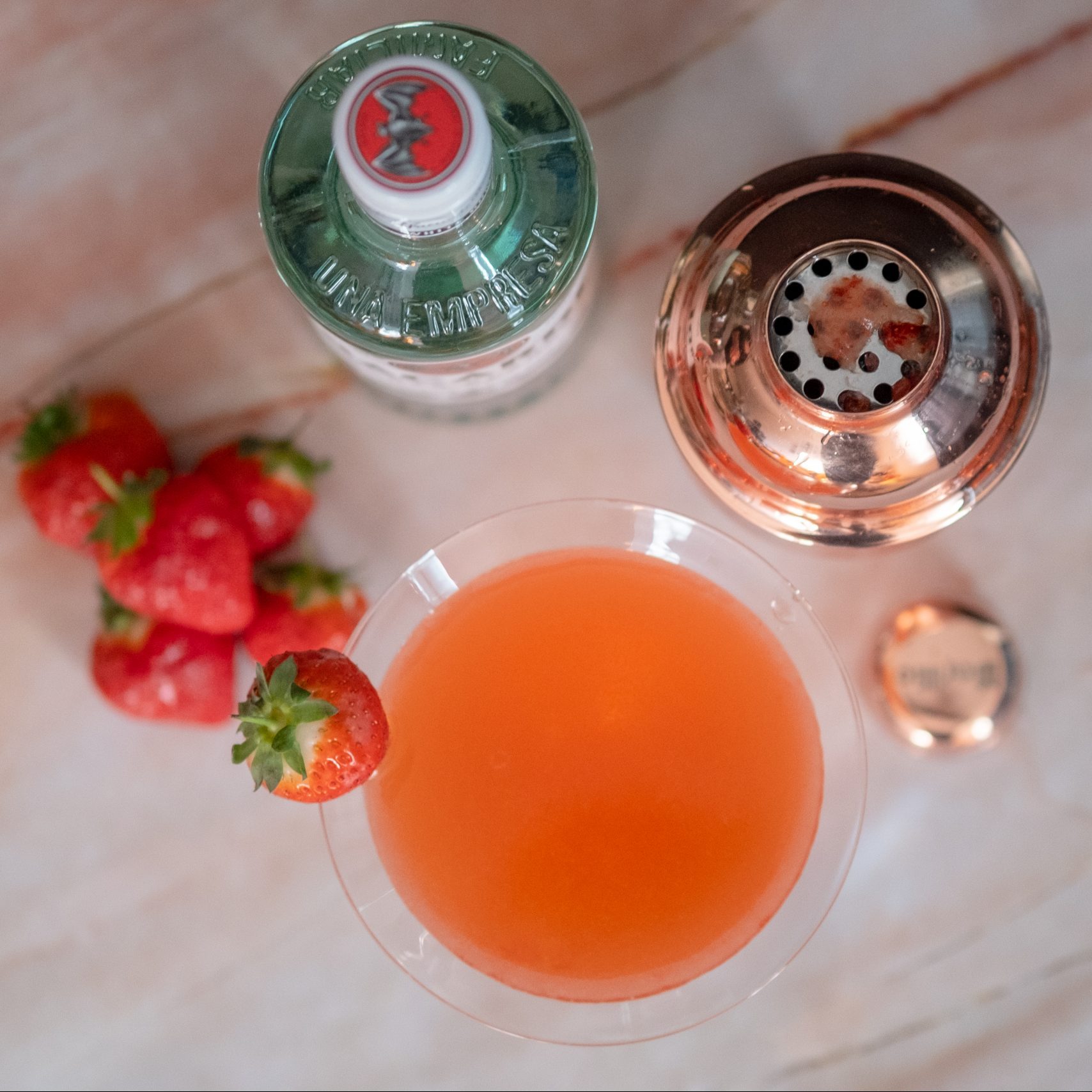 A strawberry daiquiri on a pinkish marble table with a bottle of bacardi, some strawberries and a shaker