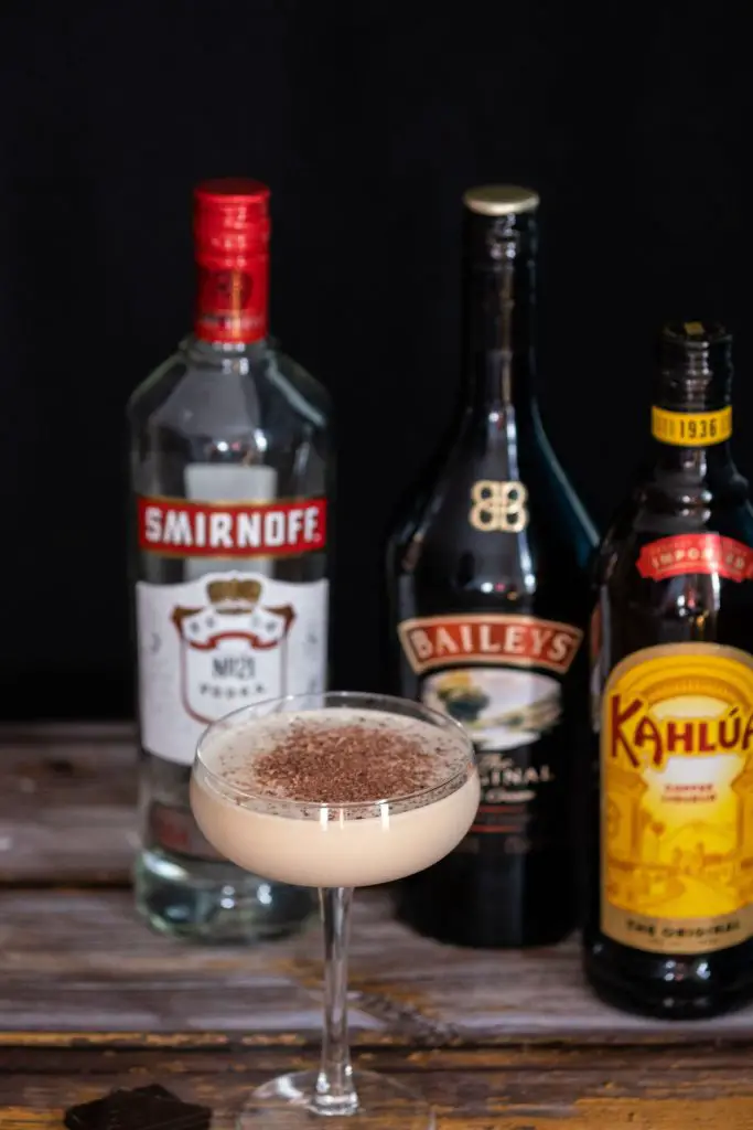 a glass of mudslide cocktail on a wooden table, with bottles of Smirnoff vodka, baileys irish cream and kahlua coffee liqueur.