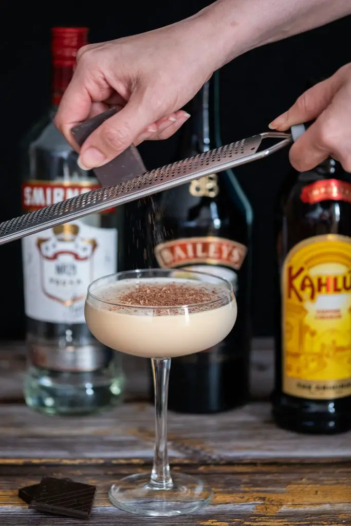 A woman hand shredding chocolate on a Mudslide cocktail from a cocktail shaker to a coupe glass on a wooden table with liqueur bottles seen blurry on the background.