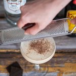 A woman hand shredding chocolate on a Mudslide cocktail from a cocktail shaker to a coupe glass on a wooden table.