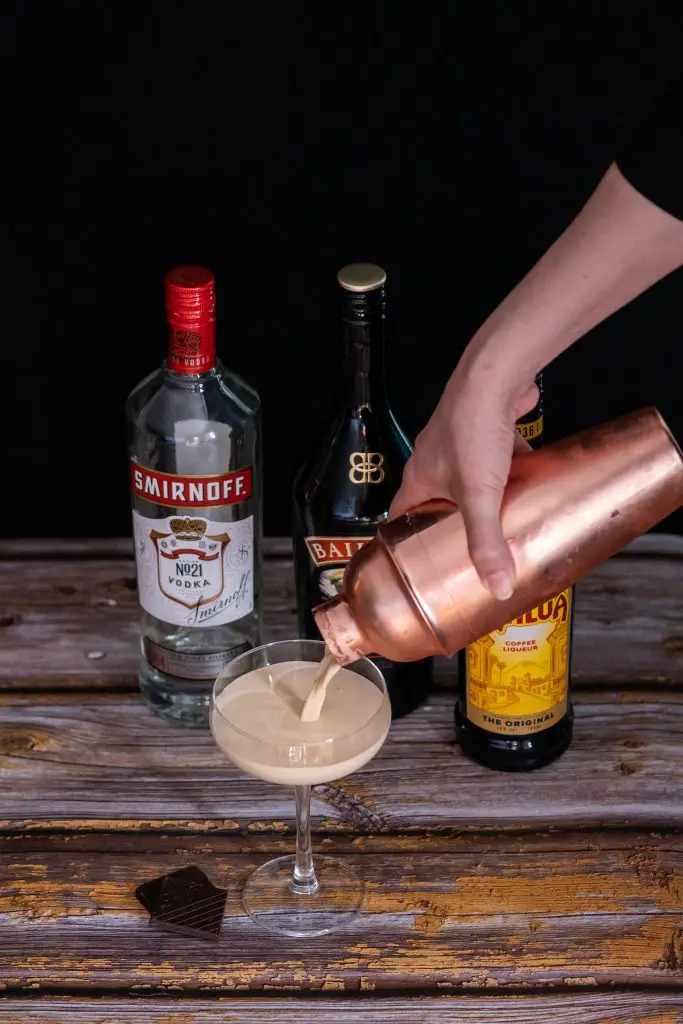 A woman hand pouring Mudslide from a cocktail shaker to a coupe glass on a wooden table with liqueur bottles seen blurry on the background.