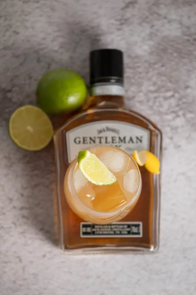 A bottle of whiskey lying on a concrete like table and a maple whiskey sour cocktail on it. There are limes around.