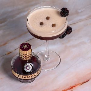 Chambord raspberry espresso martini garnished with a blackberry and coffee beans on a pink marble table, there's a bottle of chambord next to it.
