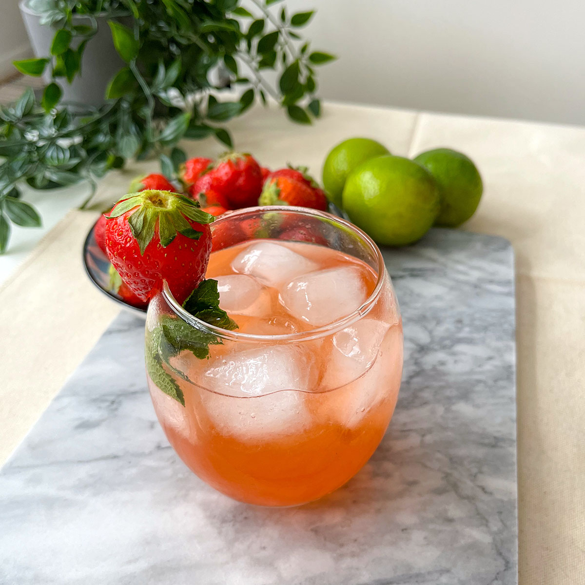 a glass of strawberry gin cocktail in a round glass filled with ice and garnished with a strawberry, sitting on a marble plate, some other strawberries and limes are seen on the background.