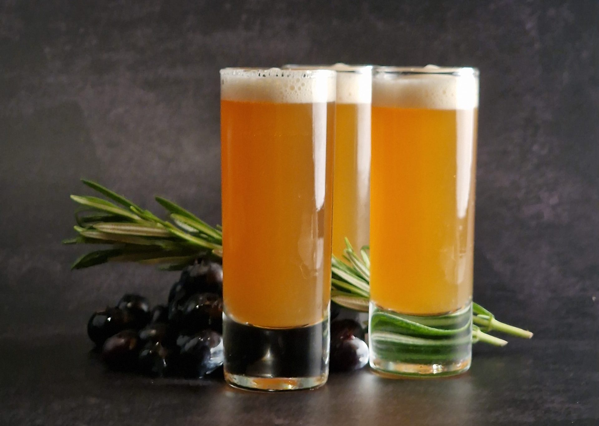 pineapple rum sherry christmas cocktail shot in 3 slim glasses with rosemary springs and some blueberries on a dark background, there's a thick frothy layer on top of the glasses. front view