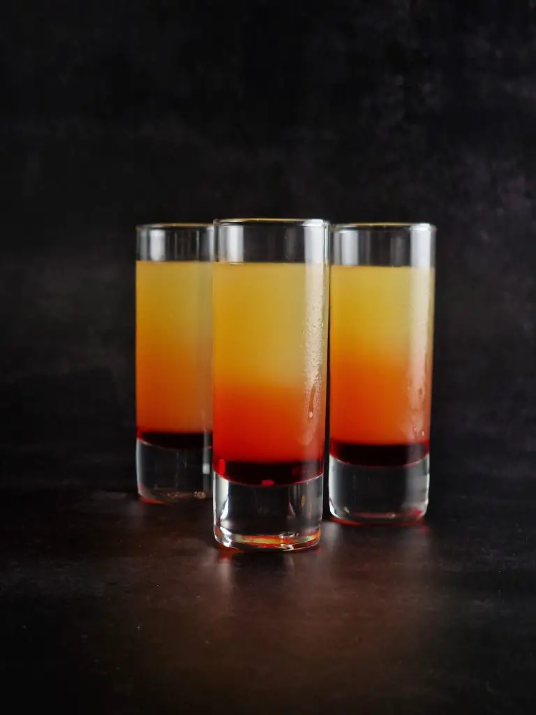 3 slim glasses front view of easy christmas cocktail shot layered from bottom to top, from red to orange and yellow colours.
