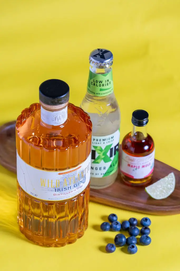 A bottle of Wild Atlantic Distillery's Irish Gin, a bottle of Lixir Ginger Ale, a small bottle of maple mule cocktail syrup, a slice of lime and some blueberries on a yellow background, 45 degrees high level angle.