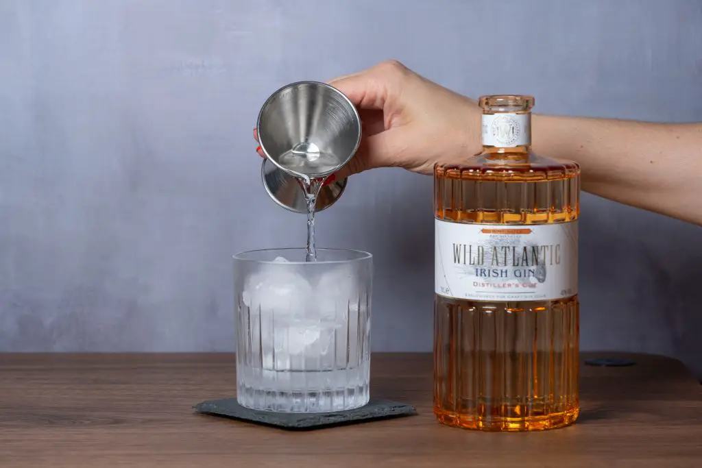 A woman's hand pouring Wild Atlantic Distillery gin with a stainless stell jigger into an old fashioned glass filled with ice to make a maple mule cocktail.