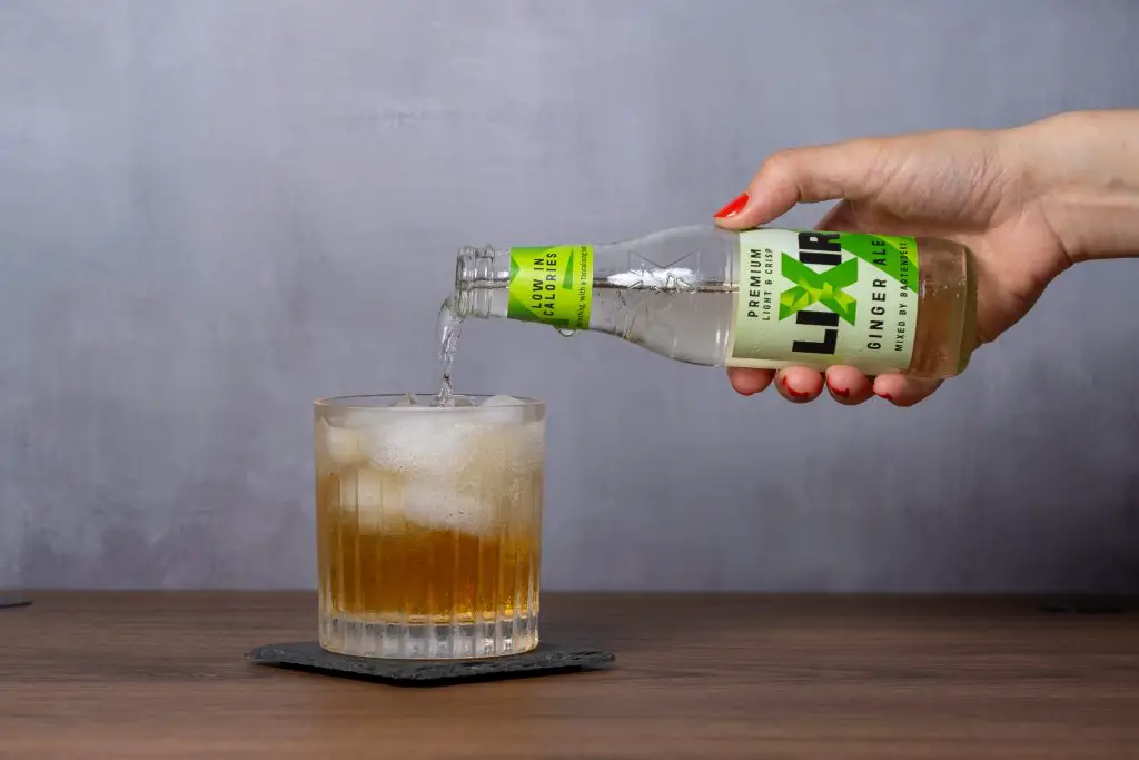 A woman's hand pouring Lixir ginger ale into an old fashioned glass to make maple mule cocktail.