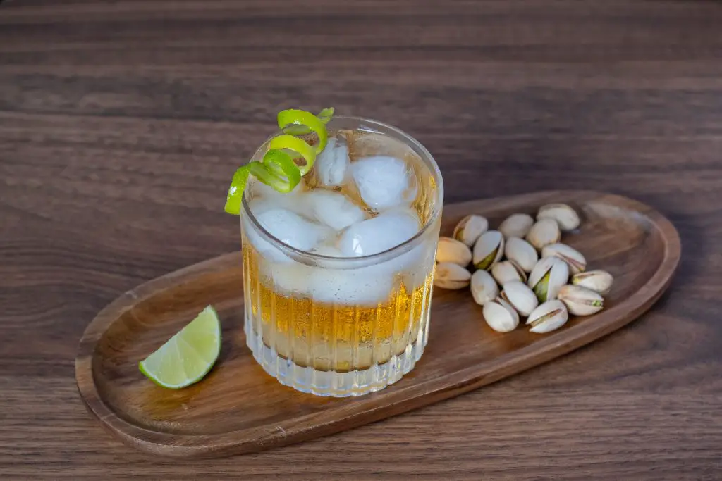 A glass of maple mule cocktail in an old fashioned glass, filled with ice and garnished with a lime peel, on a wooden tray with a lime slice and some pistachios, on a wooden table.