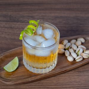 A glass of maple mule cocktail in an old fashioned glass, filled with ice and garnished with a lime peel, on a wooden tray with a lime slice and some pistachios, on a wooden table.
