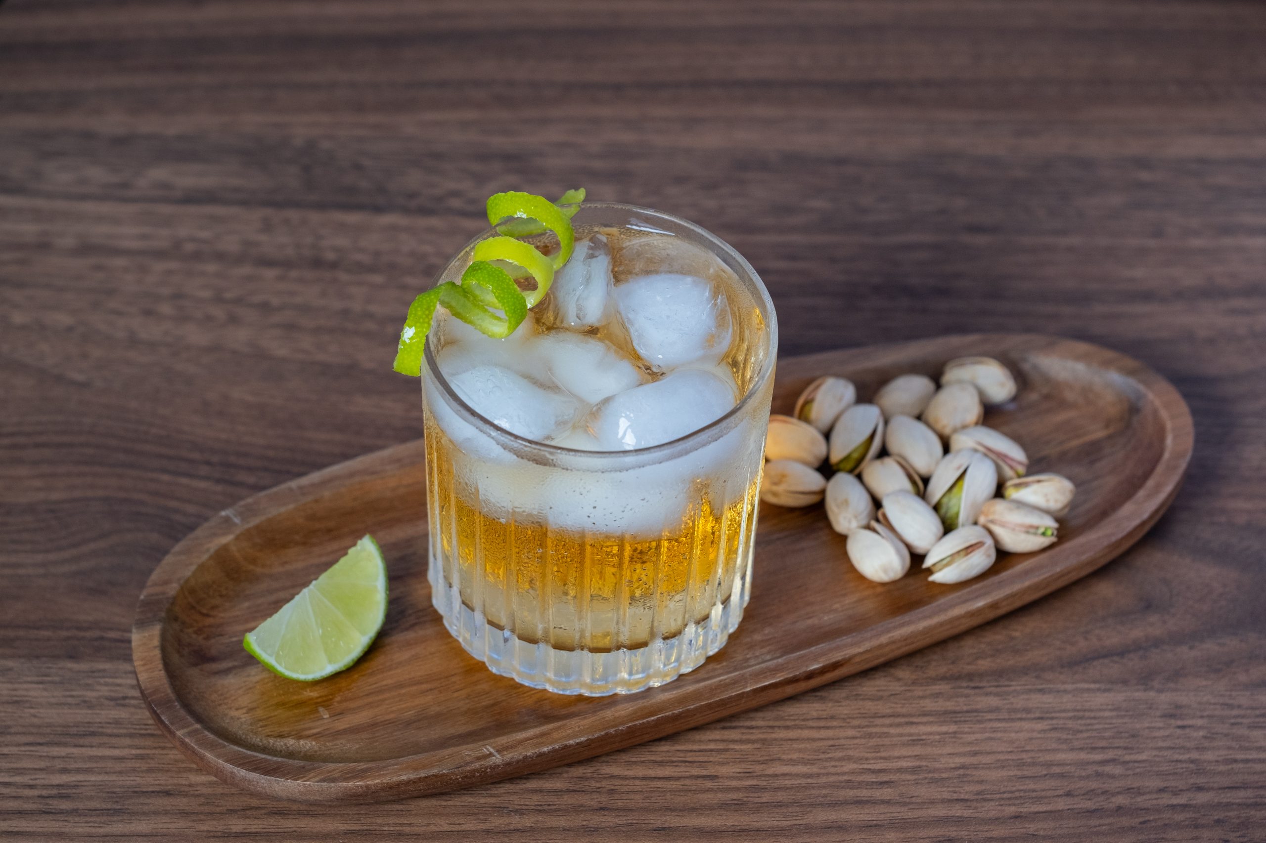 A glass of maple mule cocktail in an old fashioned glass, filled with ice and garnished with a lime peel, on a wooden tray with a lime slice and some pistachios, on a wooden table.