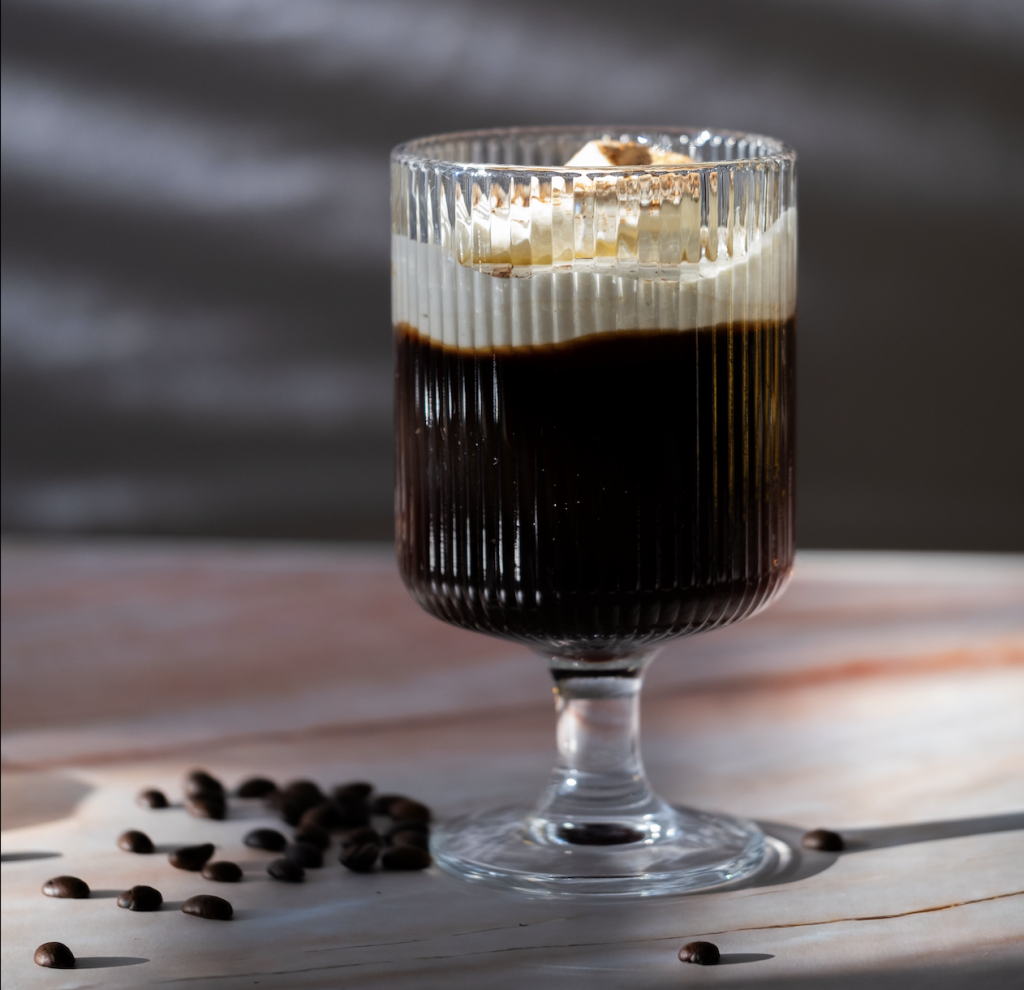 irish coffee cocktail on a pink marble table with some coffee beans spread on the table. front view square image.